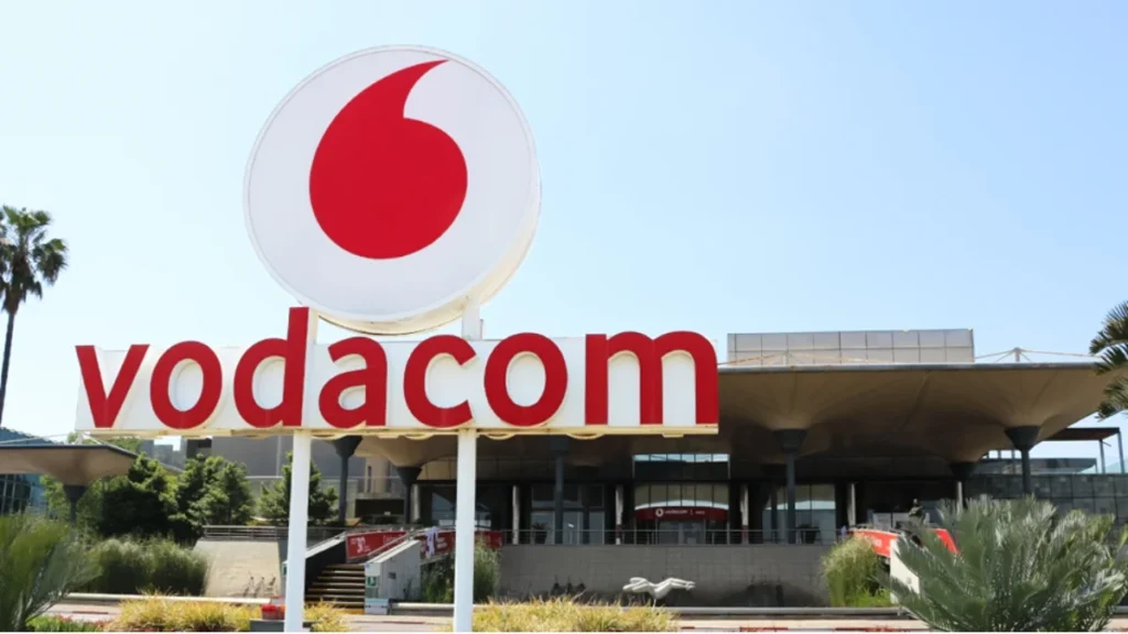 Exterior view of the Vodacom building featuring a large red logo and sign, set against a clear blue sky and palm trees.