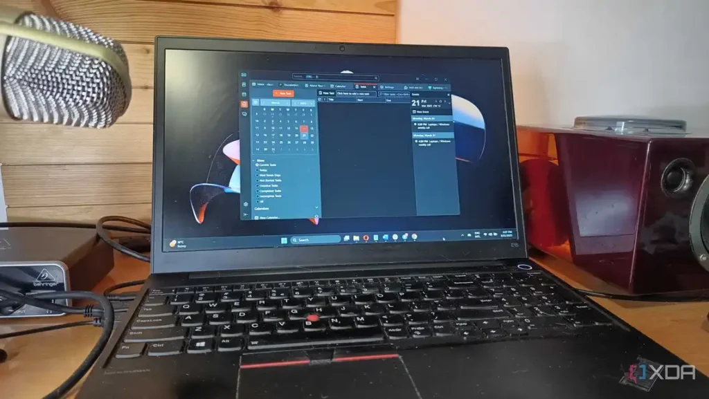 A laptop displaying a task calendar on the screen, with a microphone and speakers visible on a wooden desk.
