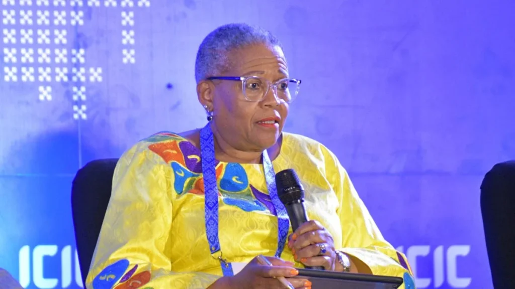 A female participant in a vibrant yellow outfit with colorful patterns speaks into a microphone at a conference, seated against a blue backdrop.