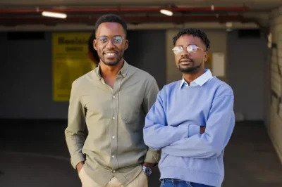 Two young men stand side by side in a parking garage, one in a green shirt and the other in a light blue sweater, exuding a casual vibe.