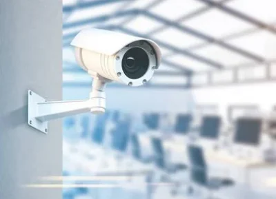 A security camera mounted on a wall, monitoring a blurred office space filled with computers and workstations.