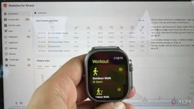 A hand holding a smartwatch displaying a workout menu, with a computer screen showing Strava activity statistics.