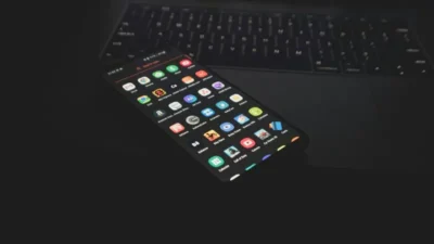 With Waydroid, It Is Easy to Run Android Apps on Linux Now 7 A smartphone displaying various app icons, resting on a dark background next to a laptop keyboard, illuminated in low light.