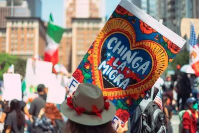 A person holds a colorful sign reading "CHINGA LA MIGRA," surrounded by a crowd at a vibrant street protest celebrating immigrant rights.