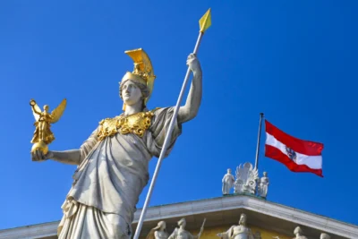 A majestic statue of a goddess holds a spear, adorned with a golden crown and robes, against a bright blue sky and Austrian flag.
