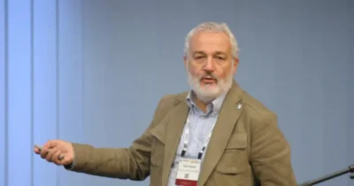 A speaker gestures while presenting in front of an audience, wearing a beige jacket and a name badge. The background is a soft blue. Featured is The Document Foundation co-founder Italo Vignoli.