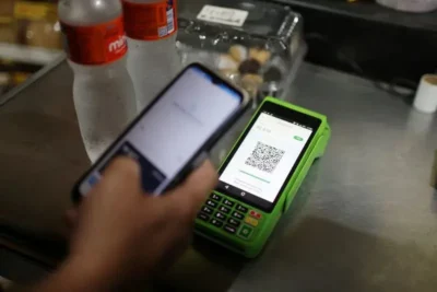 Pix payments happen in 24/7 real-time, are free, and supported by the Central Bank of Brazil 2 A hand holds a smartphone near a green payment terminal displaying a QR code, with two water bottles and pastries in the background.