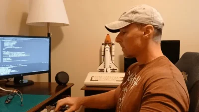 A person types on a keyboard in front of a computer screen displaying code, with a vintage computer and a model space shuttle nearby.