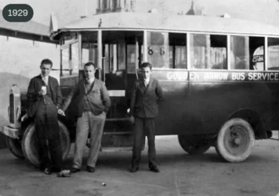 Two men in early 20th-century attire stand beside a vintage bus labeled "Gouden Arrow Bus Service" in 1929.