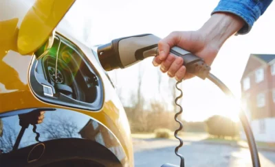 A person connects an electric vehicle charging cable to a yellow car, with sunlight glowing in the background.