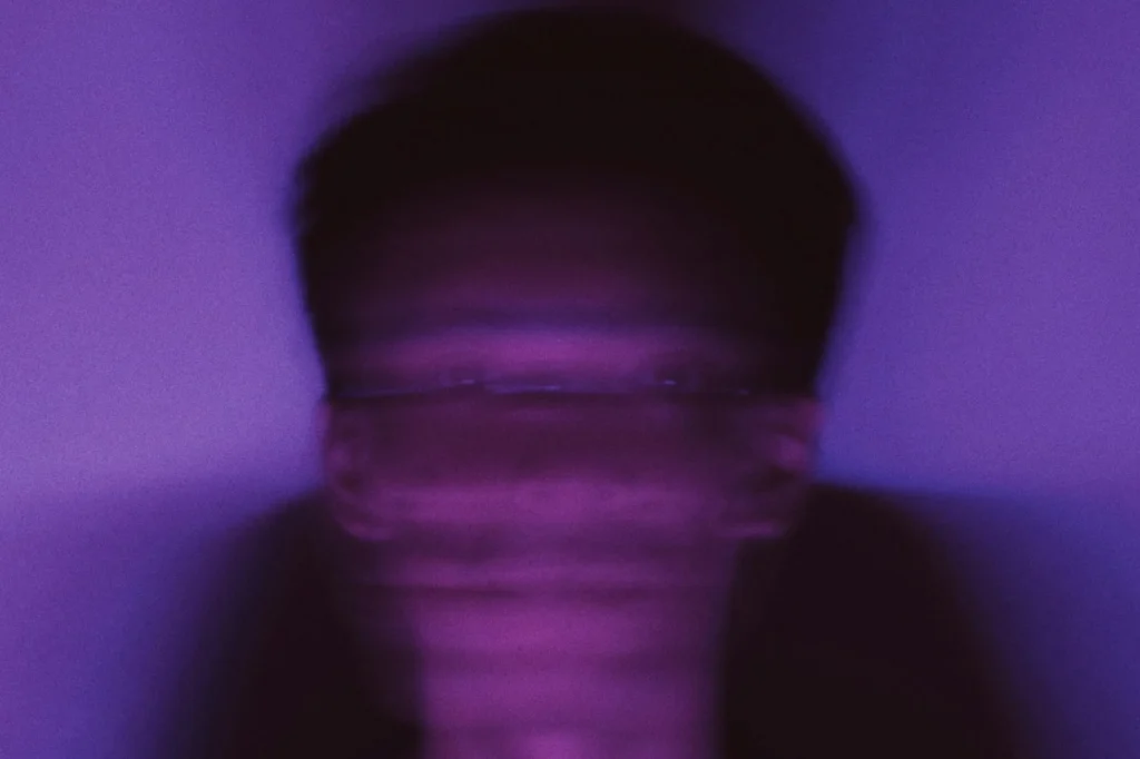 A person with dark hair in a dimly lit setting, surrounded by soft purple hues creating an atmospheric ambiance.