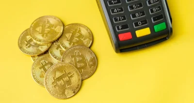A pile of gold Bitcoin coins next to a digital payment terminal on a bright yellow background.