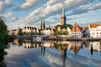 Historic buildings with colorful facades line a river, reflecting twin spires under a cloudy blue sky in Lübeck, Germany.