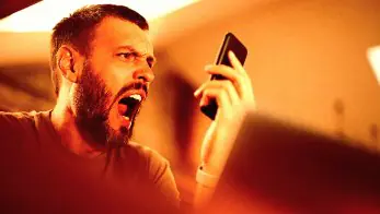 A person holds a smartphone, surrounded by warm, soft lighting that creates an intimate atmosphere. The person is shouting at their phone.