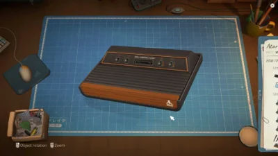 A vintage Atari video game console displayed on a blue grid mat, surrounded by tools and a mouse on a wooden desk.