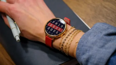 A close-up of a wrist with a gold watch featuring a digital display, surrounded by gold bracelets, with a pen resting nearby on a dark notebook.