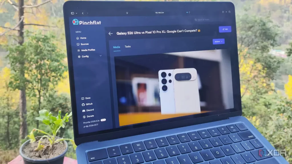 Laptop displaying a Pinchflat webpage comparing Galaxy S26 Ultra and Pixel 10 Pro XL, with a small potted plant and forest backdrop.