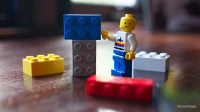 A LEGO figure with a white shirt and blue pants stands next to stacked blue and gray LEGO bricks on a wooden surface.