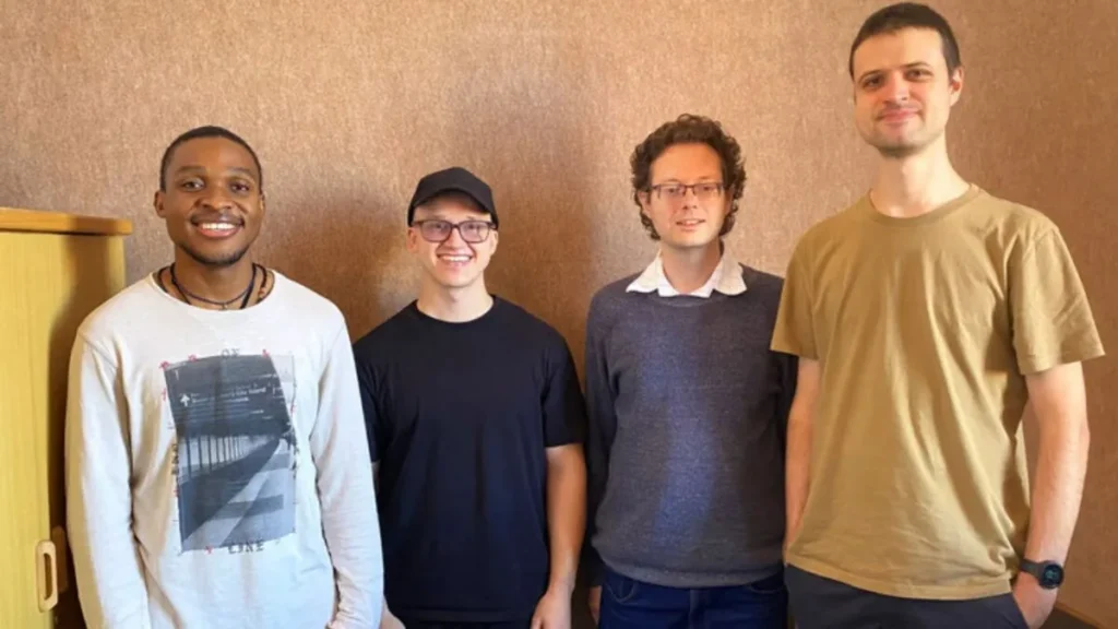 Four men standing side by side indoors against a plain wall, wearing casual clothing including t-shirts, a sweater, and a cap.