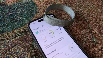 Why Bevel Is a Way Better iOS App for the Fitbit Air Than Google Health 3 Fitness tracker band resting on a smartphone displaying health stats including strain, recovery, sleep, and stress levels on a multicolored carpet.