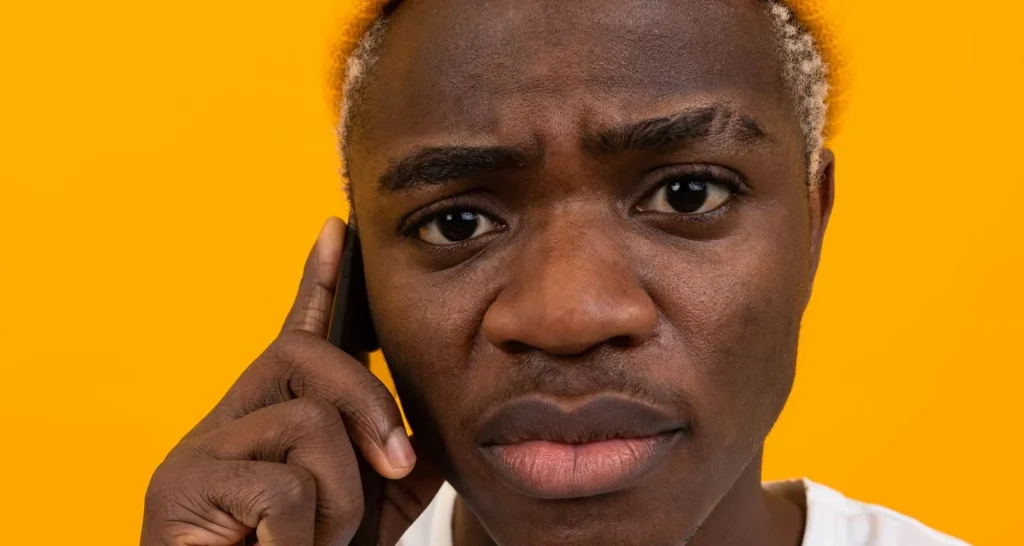 Close-up of a concerned young man with blonde hair talking on a phone against a bright orange background.