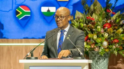 Spam callers face penalties of up to R1,000,000 in South Africa 3 Man in suit and glasses speaking at podium with microphones, floral arrangement, and map with South African and Lesotho flags in background.