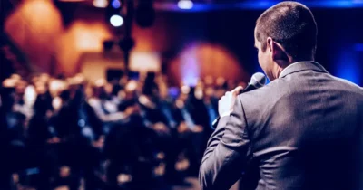 Scientist learns in front of peers why we cannot trust AI Blindly 9 Man in suit speaking into a microphone at a conference with a blurred audience in the background.