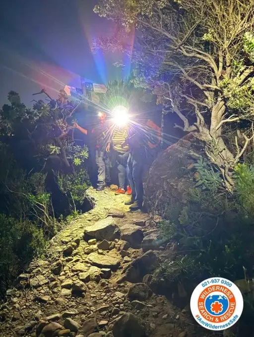 Rescue team members use headlamps while navigating a rocky, wooded mountain trail at night.